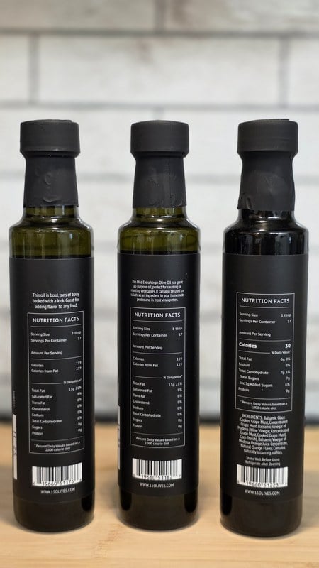 Three 15 Olives bottles on their sides showing back labels and Nutrition Facts panels, with tasting notes describing the Mild as a great all-purpose oil and the Bold as having tons of body backed with a kick