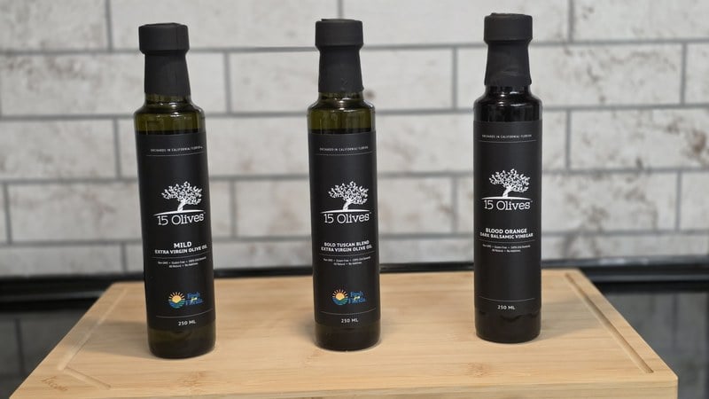 Three 15 Olives bottles standing on a bamboo cutting board against a white subway tile backsplash: Mild Extra Virgin Olive Oil, Bold Tuscan Blend Extra Virgin Olive Oil, and Blood Orange Dark Balsamic Vinegar