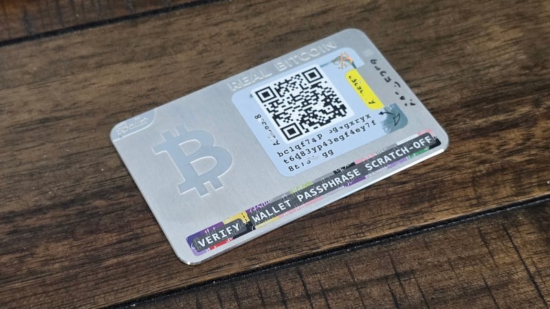 Ballet REAL stainless steel Bitcoin cold storage card on dark wood, showing front QR deposit code and scratch-off passphrase strip