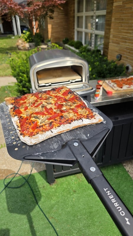 The Current Model P+ set up outdoors on a rolling cart with a finished pizza on the peel in the foreground