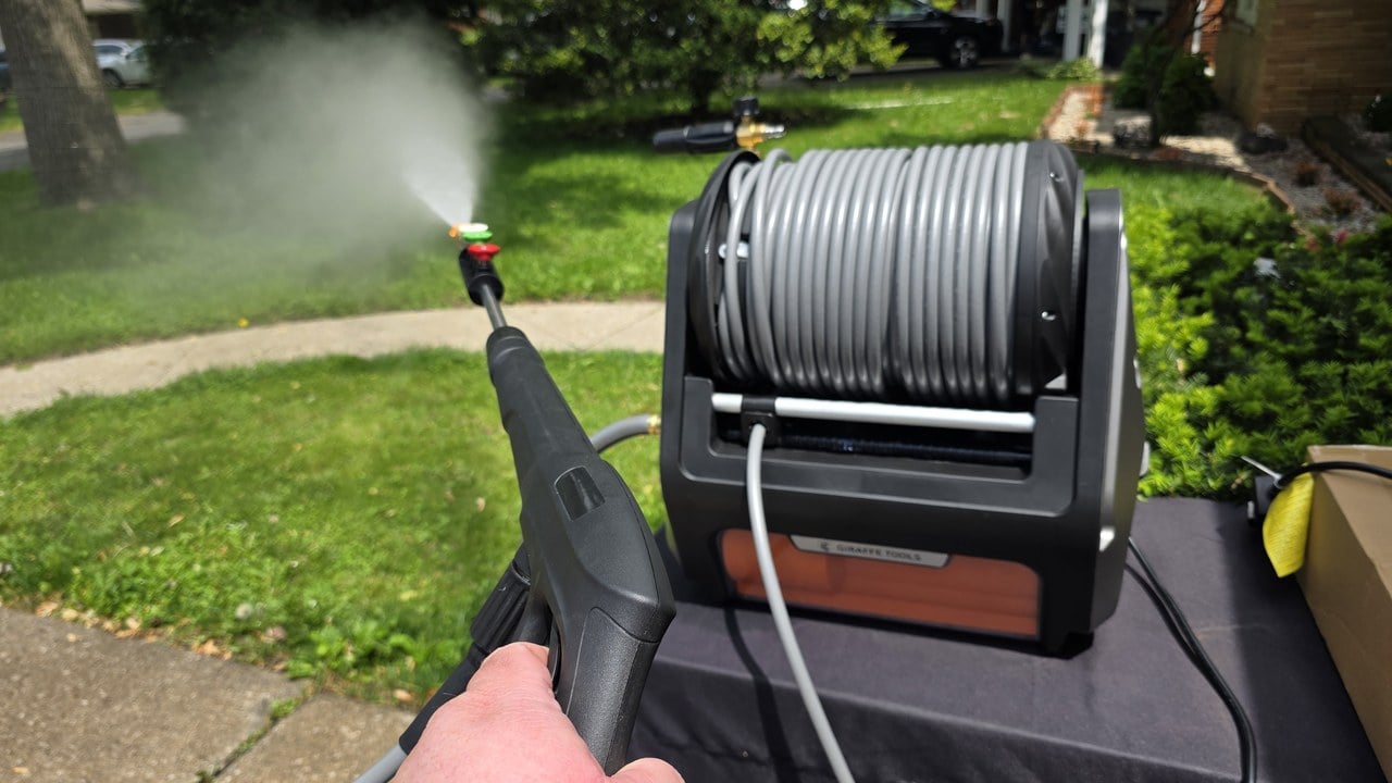 Hand pulling the trigger on the Grandfalls Pressure Washer Pro spray wand with the wall-mountable retractable hose reel beside it on a suburban driveway