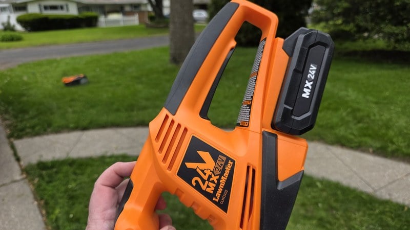 LawnMaster 24V MX battery slotted into the CLHT2422 hedge trimmer handle with a second battery visible on the lawn in the background