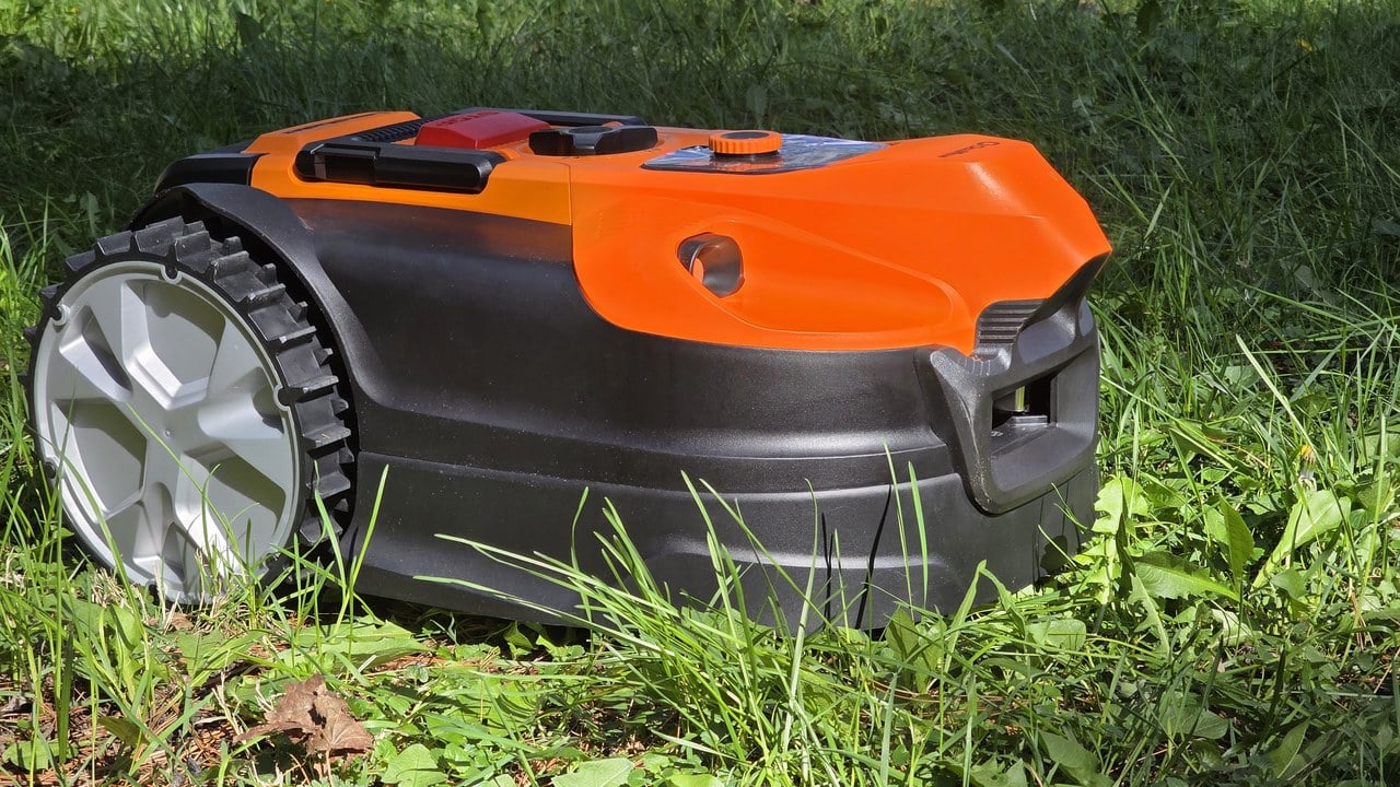LawnMaster OcuMow R1600 robot lawn mower side profile in tall sunny grass, showing orange shell and chunky knobby drive wheel