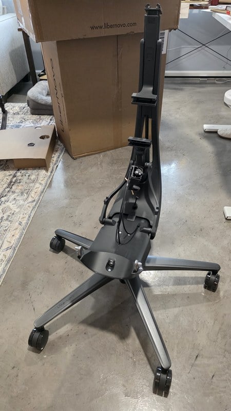 The aluminum five-star base and seat frame of the LiberNovo Maxis standing on a concrete floor during assembly, with the shipping box behind it