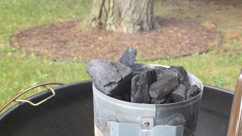 Chimney starter loaded with large chunks of hardwood lump charcoal sitting on a Weber kettle grill in a backyard
