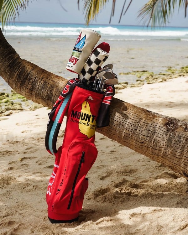 Red Mount Gay Sunday Bag with golf clubs leaning against a palm tree on a Barbados beach