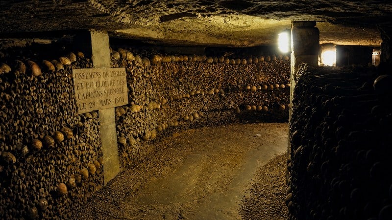 Long corridor in the Paris Catacombs lined with stacked bones and skulls, with a marble plaque visible on the left