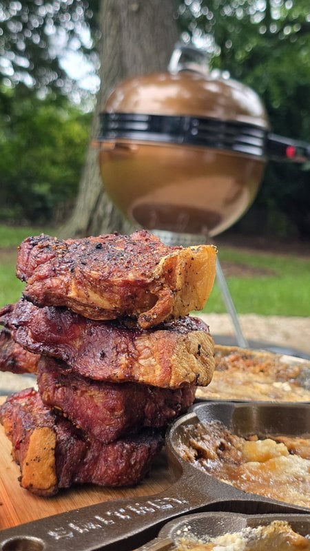 Stack of seared coppa pork steaks with crisp fat caps on a cutting board beside a cast iron pan of bourbon peach cobbler, copper Weber kettle behind them