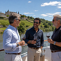 Three middle-aged men toasting with red wine on the deck of a river cruise ship passing a hilltop chateau and vineyards along a French river