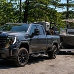 A dark gray GMC Sierra Denali HD pickup towing a center-console fishing boat on a tandem trailer at a tree-lined northern Michigan lake boat ramp