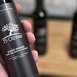 15 Olives Blood Orange Dark Balsamic Vinegar bottle held in hand, showing California and Florida orchards label, with two soft-focus 15 Olives bottles on a wooden surface in the background