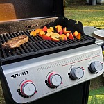 Steak and sliced peaches grilling on a Weber Spirit gas grill in a backyard, with tongs, a plate and a stainless fire pit alongside