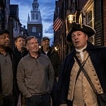 Costumed guide holding a lantern leads a group of men on a nighttime ghost tour through a historic brick alley