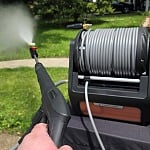 Hand pulling the trigger on the Grandfalls Pressure Washer Pro spray wand with the wall-mountable retractable hose reel beside it on a suburban driveway