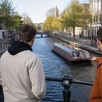 Three guys leaning on a canal bridge railing watching a tour boat pass through an Amsterdam canal