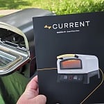 The Current Model P+ Smart Pizza Oven manual held up outdoors in front of an open grill