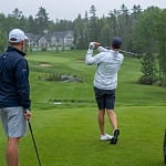 Two men golf in light rain on a tree-lined northern course, one finishing his swing on the tee box beside a cart loaded with golf bags