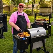 James Hills at the grill surrounded by a backyard cooking stack including a Masterbuilt Gravity Series smoker, a Z Grills pellet grill, a Santa Maria-style grill, and a stainless 4-burner gas grill