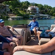 Five guys hanging out on a pontoon boat with bluff-side lake homes and covered boat slips behind them at Lake of the Ozarks