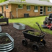Backyard spring cleanup scene with galvanized raised beds, wood planter box, dump cart full of yard debris, and red zero-turn riding mower on a freshly mulched and cleaned lawn