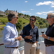Three middle-aged men toasting with red wine on the deck of a river cruise ship passing a hilltop chateau and vineyards along a French river