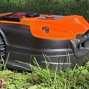 LawnMaster OcuMow R1600 robot lawn mower side profile in tall sunny grass, showing orange shell and chunky knobby drive wheel