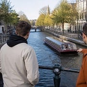 Three guys leaning on a canal bridge railing watching a tour boat pass through an Amsterdam canal