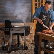 A dad in a Kiss the Cook apron slicing brisket beside a Traeger pellet grill