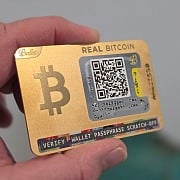 Gold-plated Ballet REAL Bitcoin cold storage wallet held in hand, showing front QR deposit address and lottery-style scratch-off passphrase strip