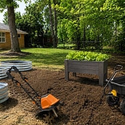 Backyard vegetable garden setup with raised garden beds, electric tiller, Step2 Lakewood planter box with lettuce, and Gorilla Carts dump cart on freshly tilled soil