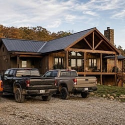 A modern timber-frame hunting cabin with a black metal roof, covered porch, and elevated deck, with three trucks parked on the gravel drive amid fall foliage in rural Tennessee.