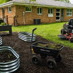 Backyard spring cleanup scene with galvanized raised beds, wood planter box, dump cart full of yard debris, and red zero-turn riding mower on a freshly mulched and cleaned lawn