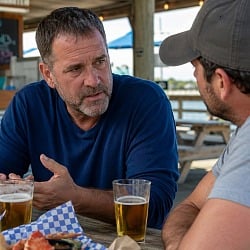 Two men talking over beers and seafood at a dockside table, one checking in on the other