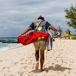 A golfer walks a Barbados beach carrying the red Mount Gay 1703 Devereux Golf bag and checkered towel