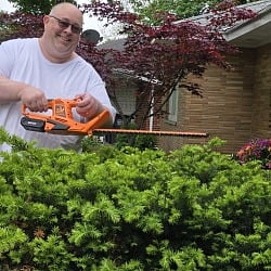 Man trimming a yew bush with the orange LawnMaster 22-Inch 24V Cordless Hedge Trimmer in front of a brick house