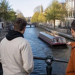 Three guys leaning on a canal bridge railing watching a tour boat pass through an Amsterdam canal