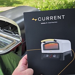 The Current Model P+ Smart Pizza Oven manual held up outdoors in front of an open grill