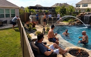 Guys hanging out around a backyard pool with a fire pit, outdoor kitchen and a pergola strung with lights