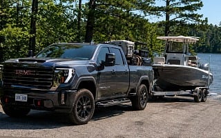 A dark gray GMC Sierra Denali HD pickup towing a center-console fishing boat on a tandem trailer at a tree-lined northern Michigan lake boat ramp