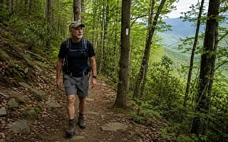 A man hiking solo on a section of the Appalachian Trail, a white blaze on a tree marking the route, green mountains beyond.