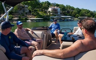 Five guys hanging out on a pontoon boat with bluff-side lake homes and covered boat slips behind them at Lake of the Ozarks