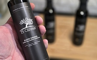 15 Olives Blood Orange Dark Balsamic Vinegar bottle held in hand, showing California and Florida orchards label, with two soft-focus 15 Olives bottles on a wooden surface in the background
