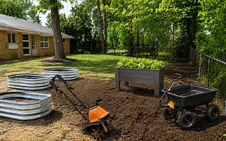Backyard vegetable garden setup with raised garden beds, electric tiller, Step2 Lakewood planter box with lettuce, and Gorilla Carts dump cart on freshly tilled soil