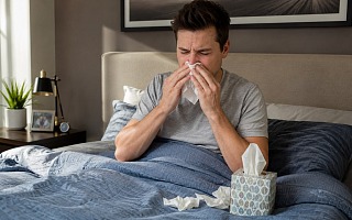 Man sitting up in bed blowing his nose into a tissue, with a tissue box on the blue bedding