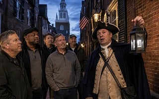 Costumed guide holding a lantern leads a group of men on a nighttime ghost tour through a historic brick alley