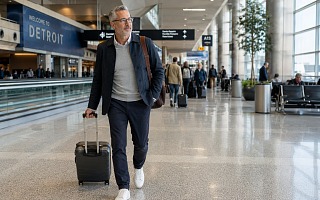 A well-dressed man with a small carry-on suitcase walking through Detroit airport's terminal in a navy jacket and sneakers