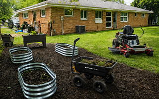 Backyard spring cleanup scene with galvanized raised beds, wood planter box, dump cart full of yard debris, and red zero-turn riding mower on a freshly mulched and cleaned lawn