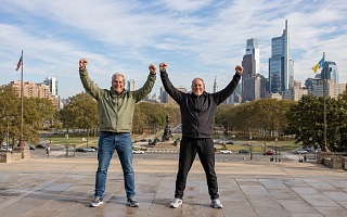 Two men at the top of the Philadelphia Museum of Art steps with their arms raised, the Benjamin Franklin Parkway and the Center City skyline behind them