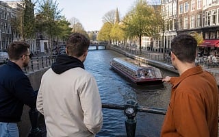 Three guys leaning on a canal bridge railing watching a tour boat pass through an Amsterdam canal