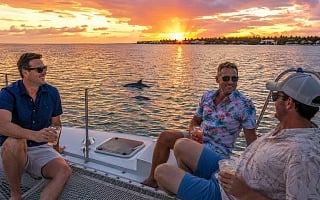 Three guys with drinks on a catamaran at sunset as dolphins surface on a Key West eco boat tour